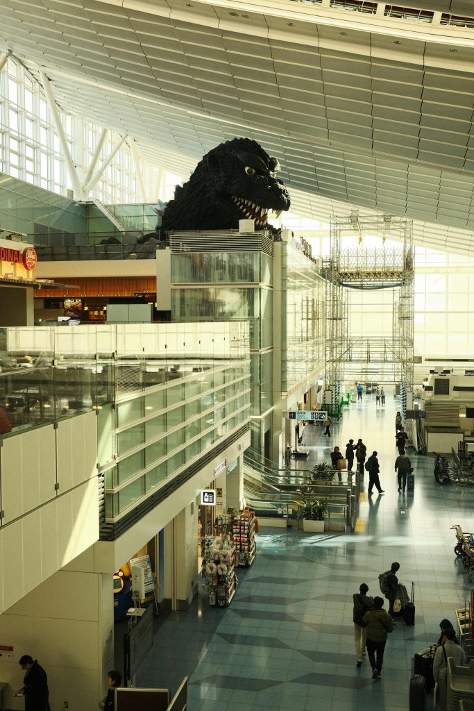 Iconic Godzilla statue inside a bustling, modern airport terminal with a high ceiling.